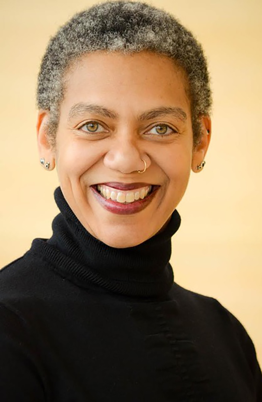 Portrait headshot photograph of Rhonda Y. Williams smiling in a black turtleneck long-sleeve shirt, wearing two chrome-colored earrings, and has a gold-colored nose piercing plus minimal slight faded dark black lipstick